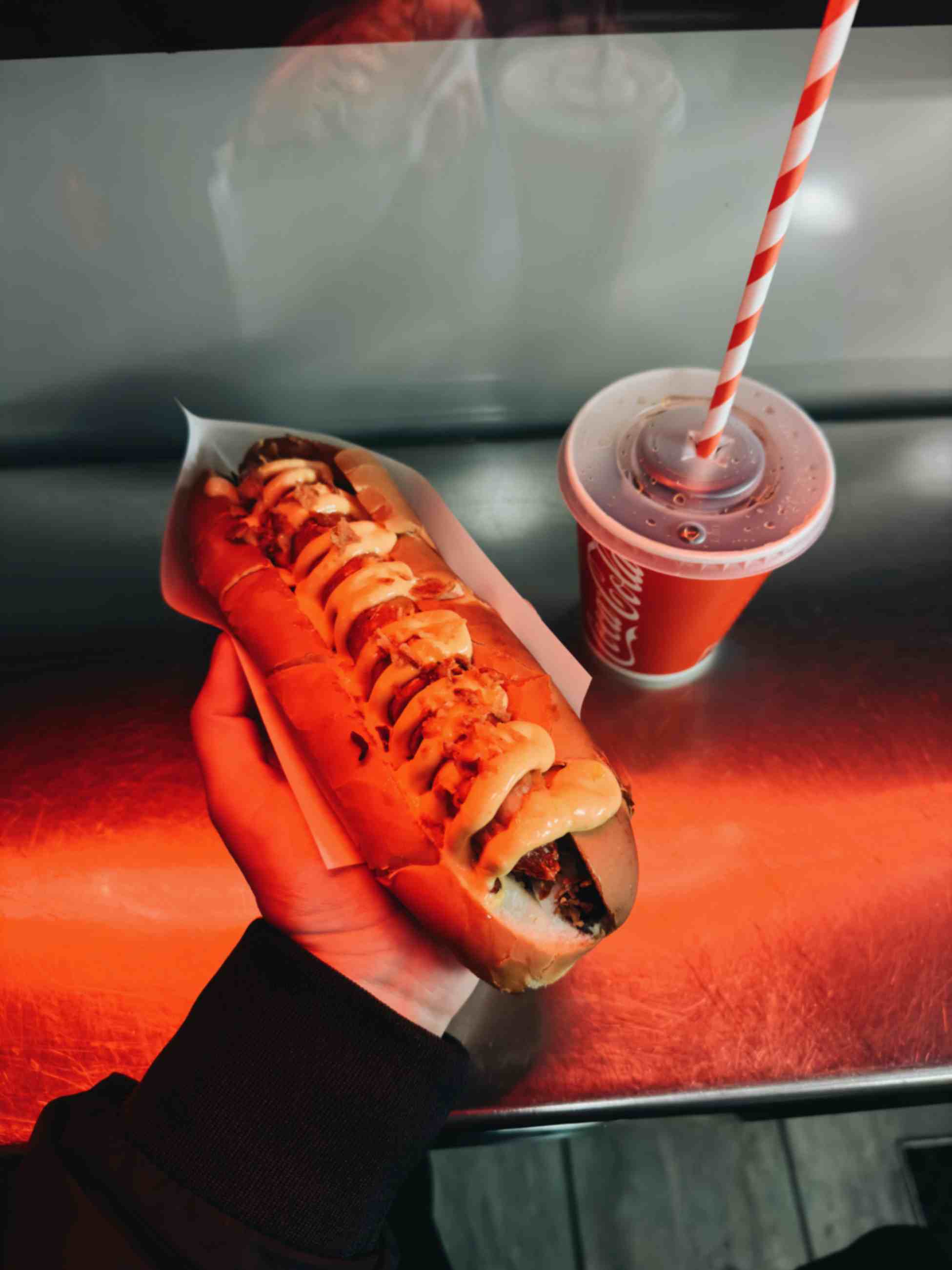 Hand holding a hot dog topped with sauce and onions next to a soft drink with a striped straw on a counter.