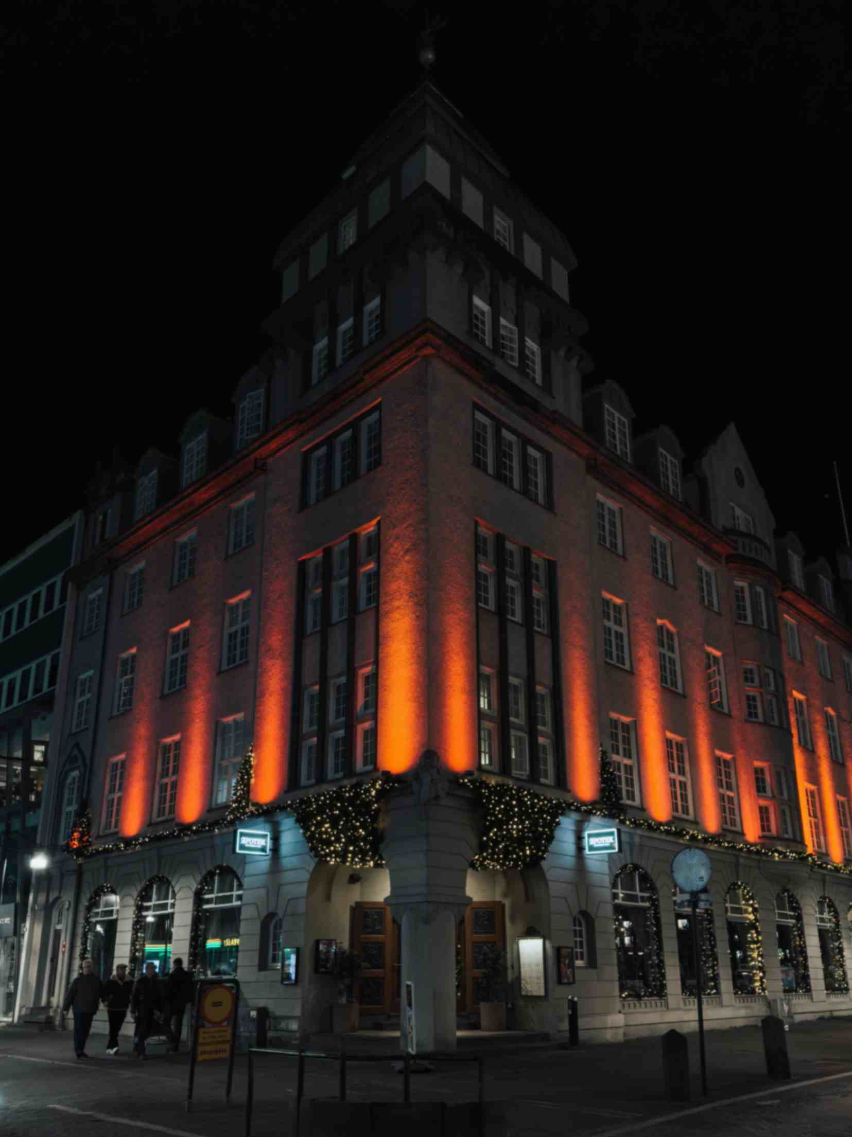 Multi-story building lit with warm orange lights at night, with people walking on the street below.
