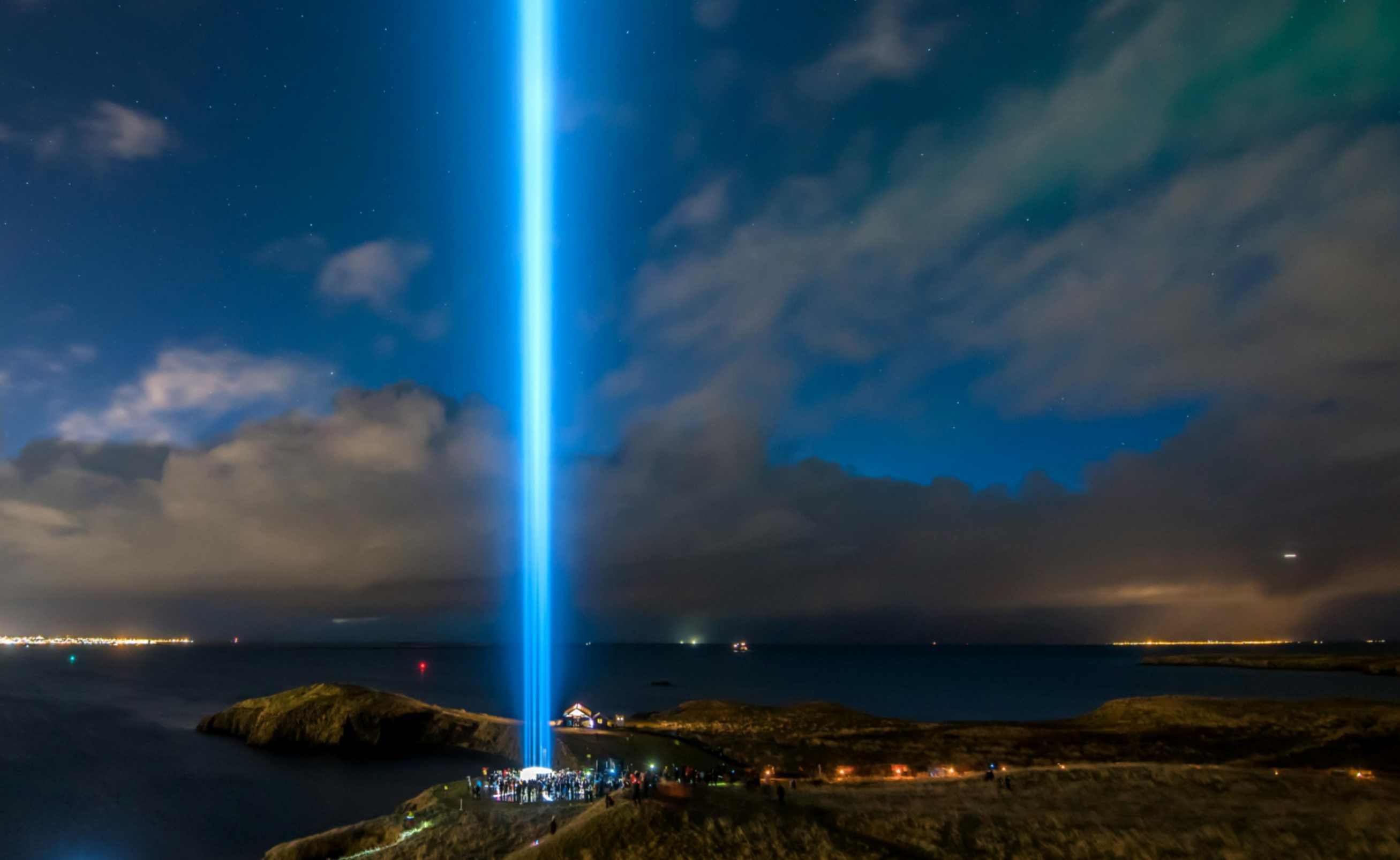 Bright vertical light beam shining into the night sky from a small island, with people gathered below near the water.