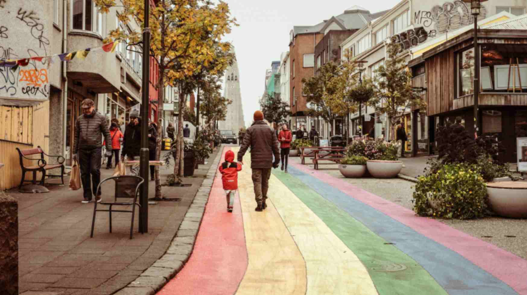 People walking along a colorful rainbow-painted street lined with shops and trees in a city center.