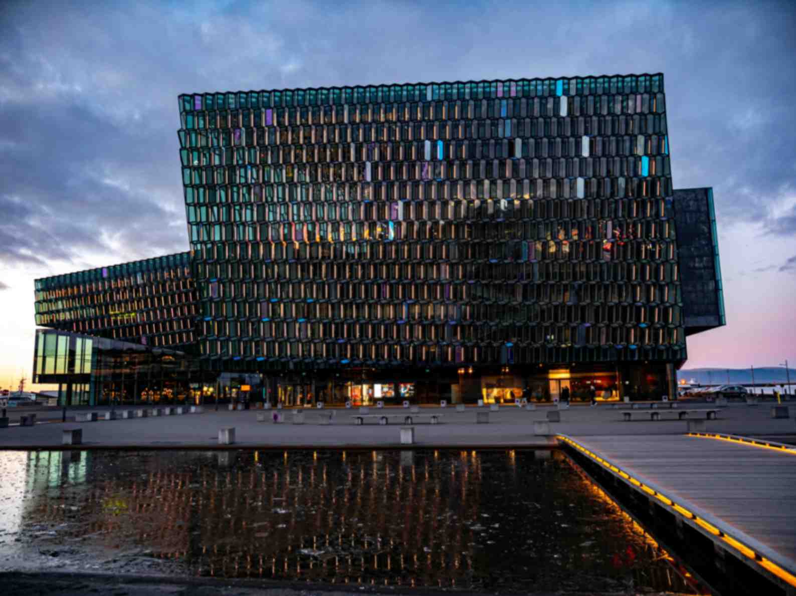 Modern glass building with a geometric facade reflecting light, situated by water at dusk.