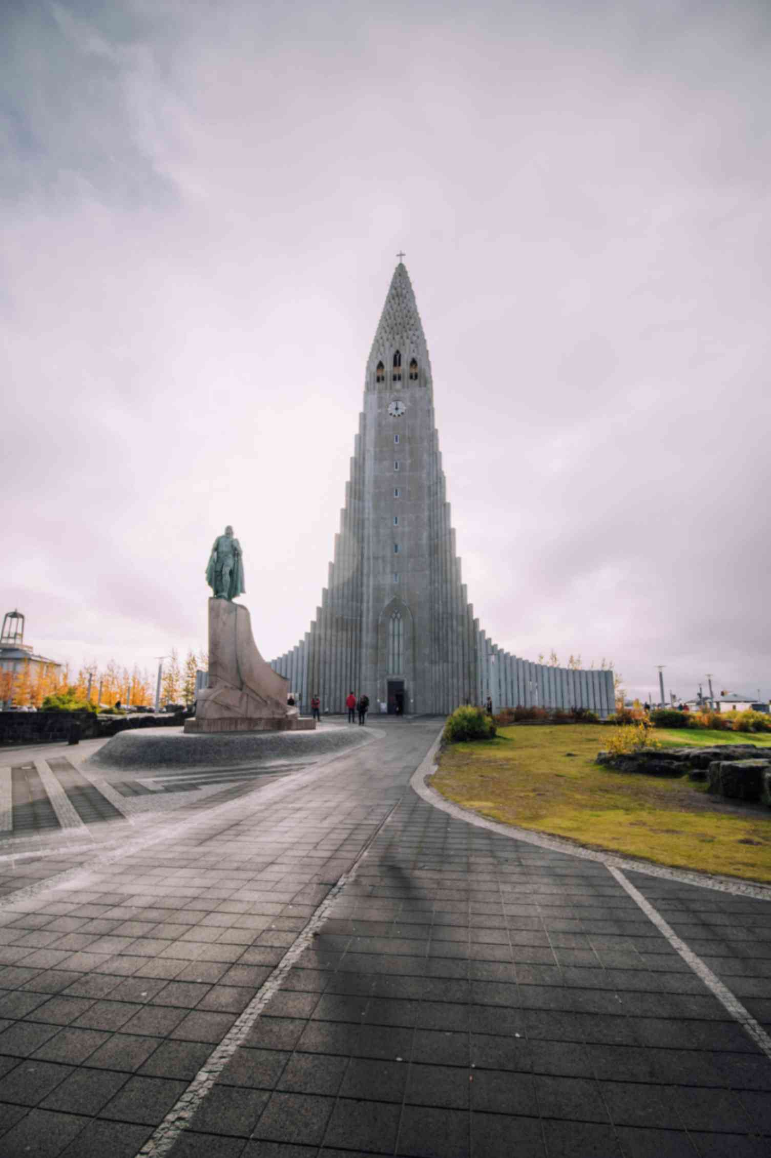 Tall gray church with a stepped tower rising above a plaza, with a statue in the foreground under a cloudy sky
