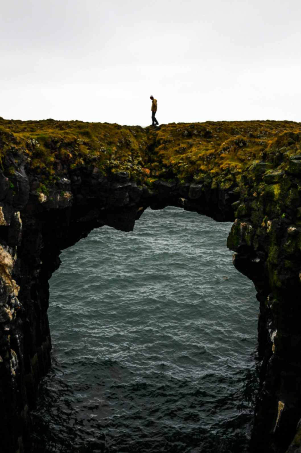 Hiker walking across natural sea arch cliff above ocean along rugged Iceland coastline