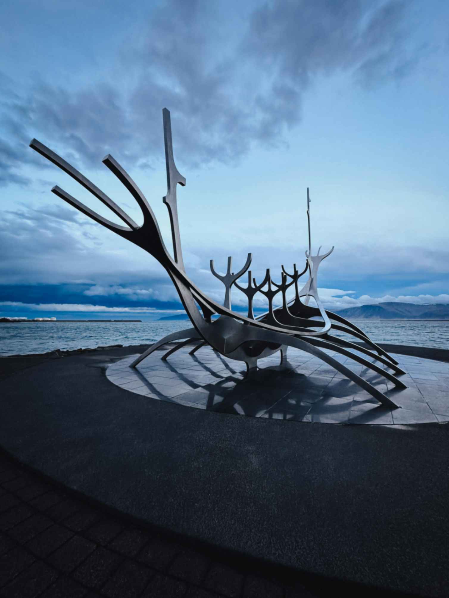 Abstract metal boat sculpture on a waterfront with ocean and mountains in the background under a cloudy sky.