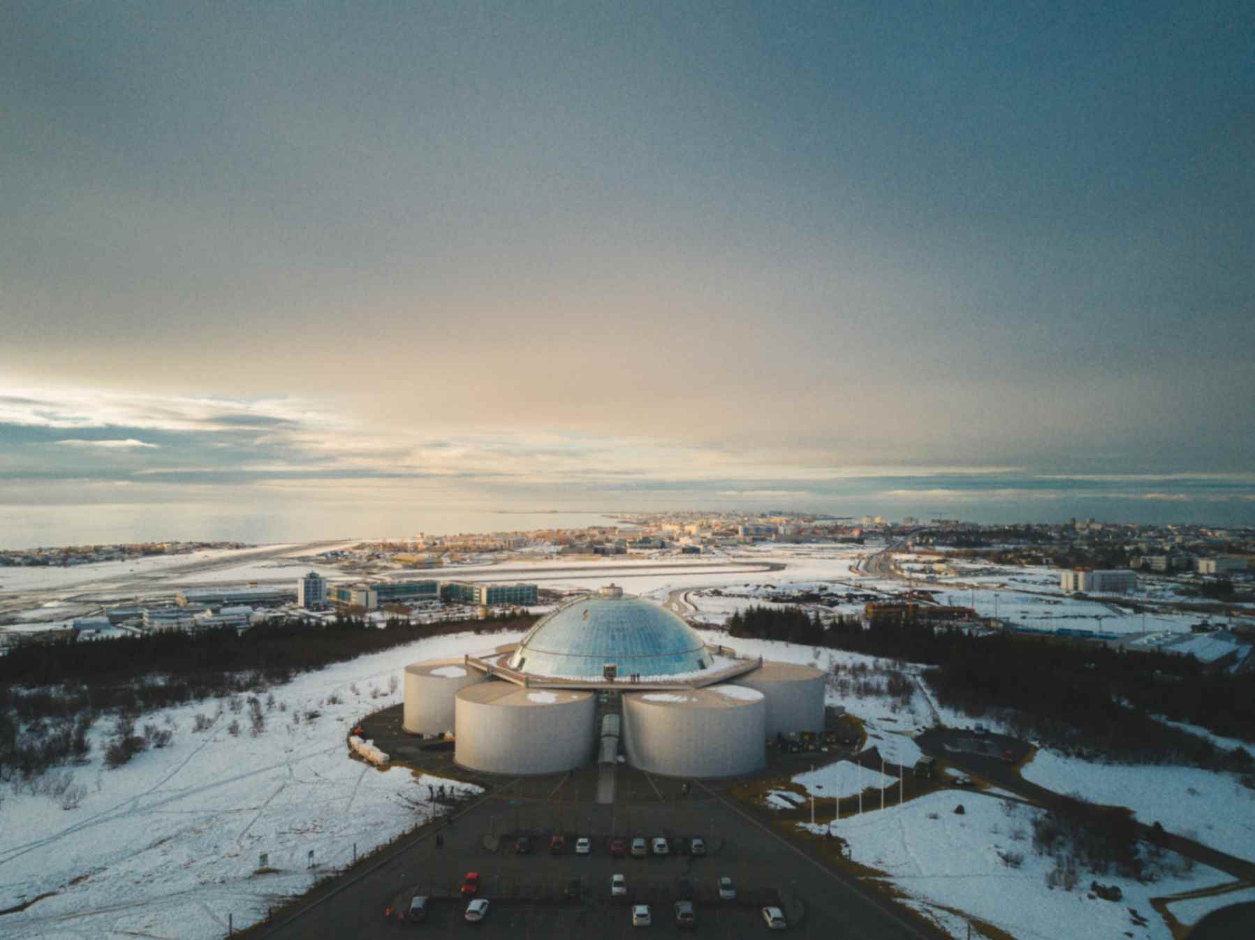 Large glass-domed building on a hill surrounded by snow with a cityscape in the background.